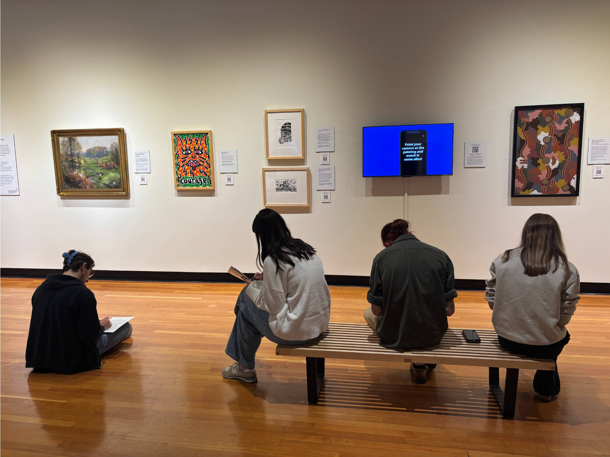 An art gallery with framed artworks on the wall and one TV displaying a video. In front of the wall sit four college students who are looking down and writing. Their backs are to the viewer.
